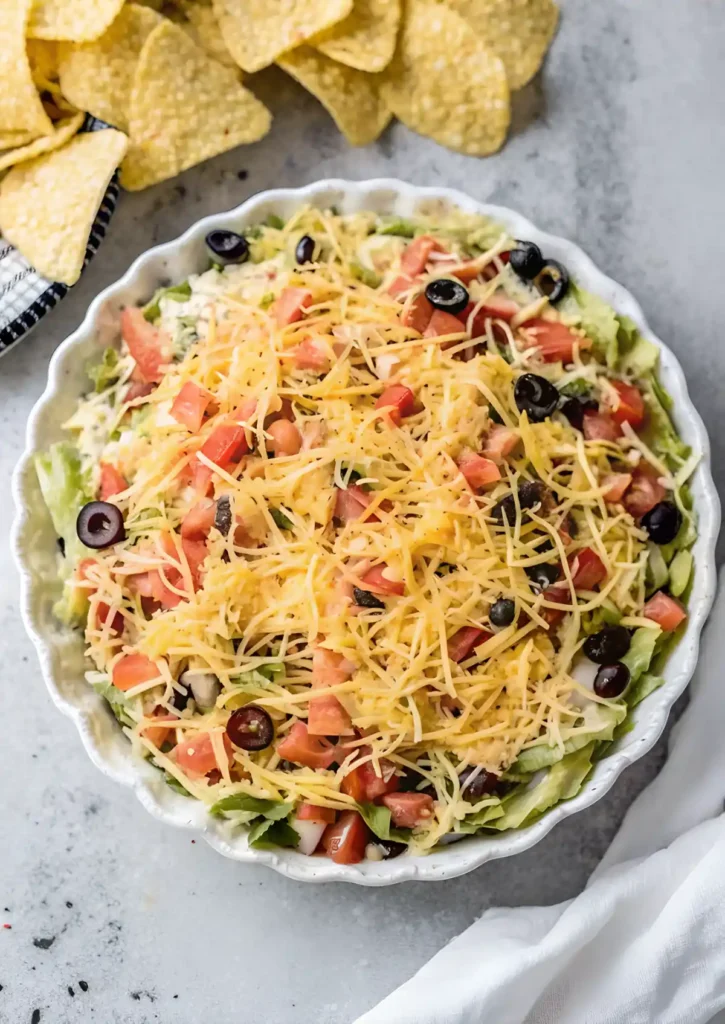Overhead view of no cook taco dip topped with shredded cheddar, diced tomatoes, black olives, and lettuce in a white scalloped dish with tortilla chips nearby.