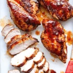 Juicy BBQ chicken breasts glazed with sauce on a cutting board, sliced to show moist interior, with a red basting brush on the side.
