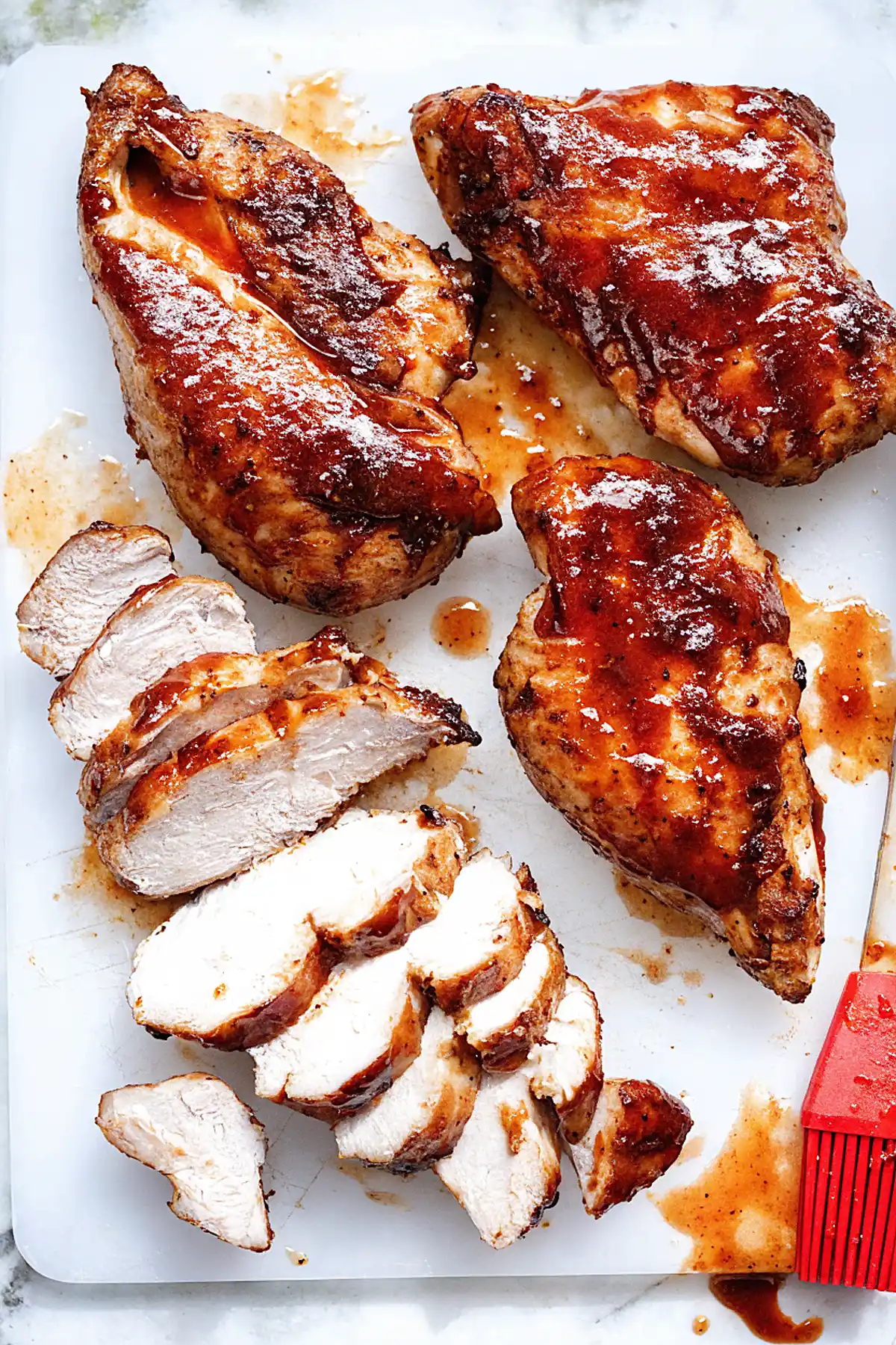 Juicy BBQ chicken breasts glazed with sauce on a cutting board, sliced to show moist interior, with a red basting brush on the side.