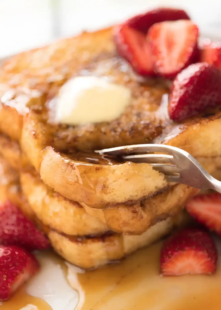 Fork lifting a soft bite of French toast with maple syrup, butter, and fresh strawberries.