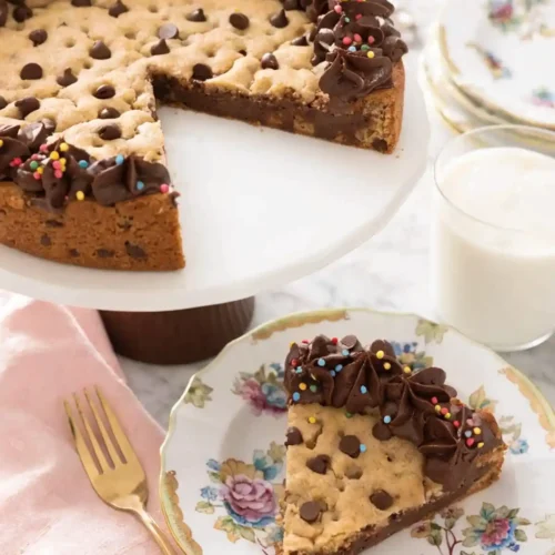 Chocolate chip cookie cake with chocolate frosting border and sprinkles on a cake stand, served with a slice on a floral plate and a glass of milk.