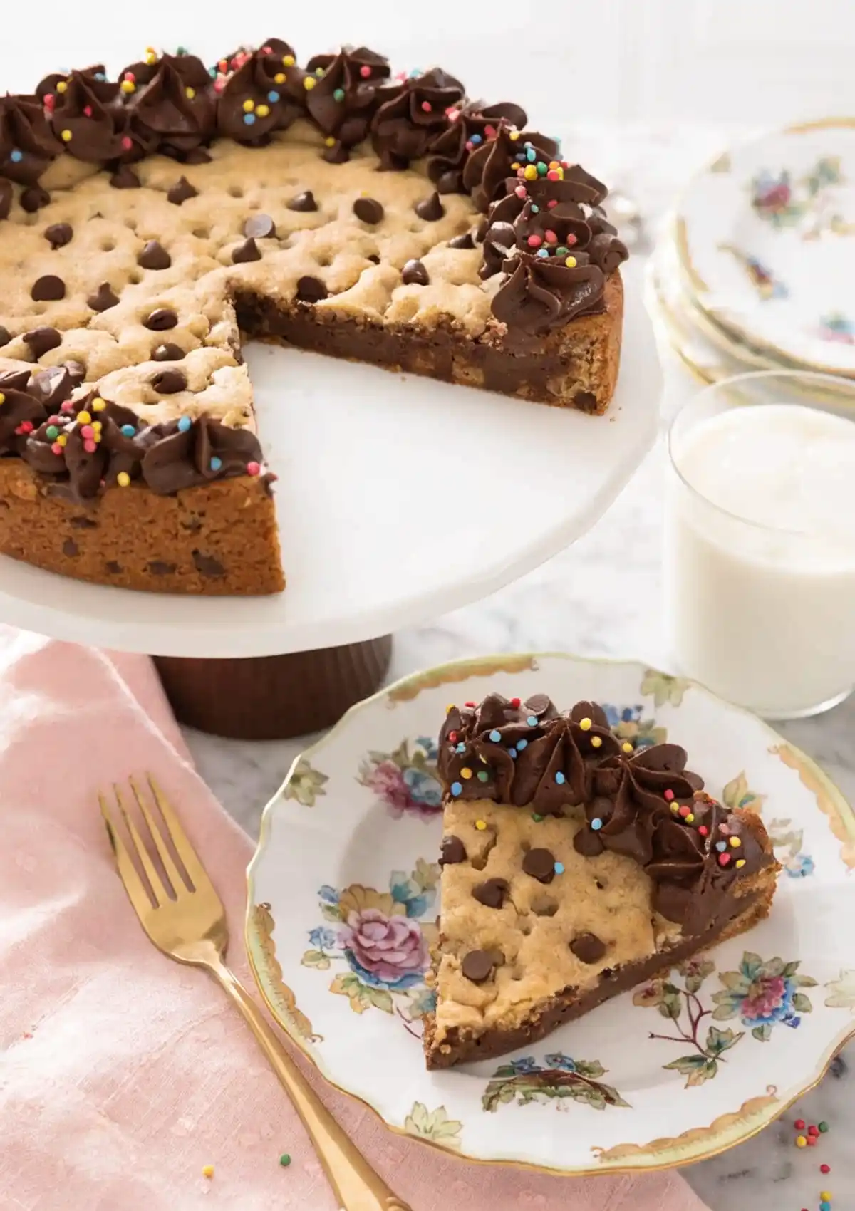 Chocolate chip cookie cake with chocolate frosting border and sprinkles on a cake stand, served with a slice on a floral plate and a glass of milk.