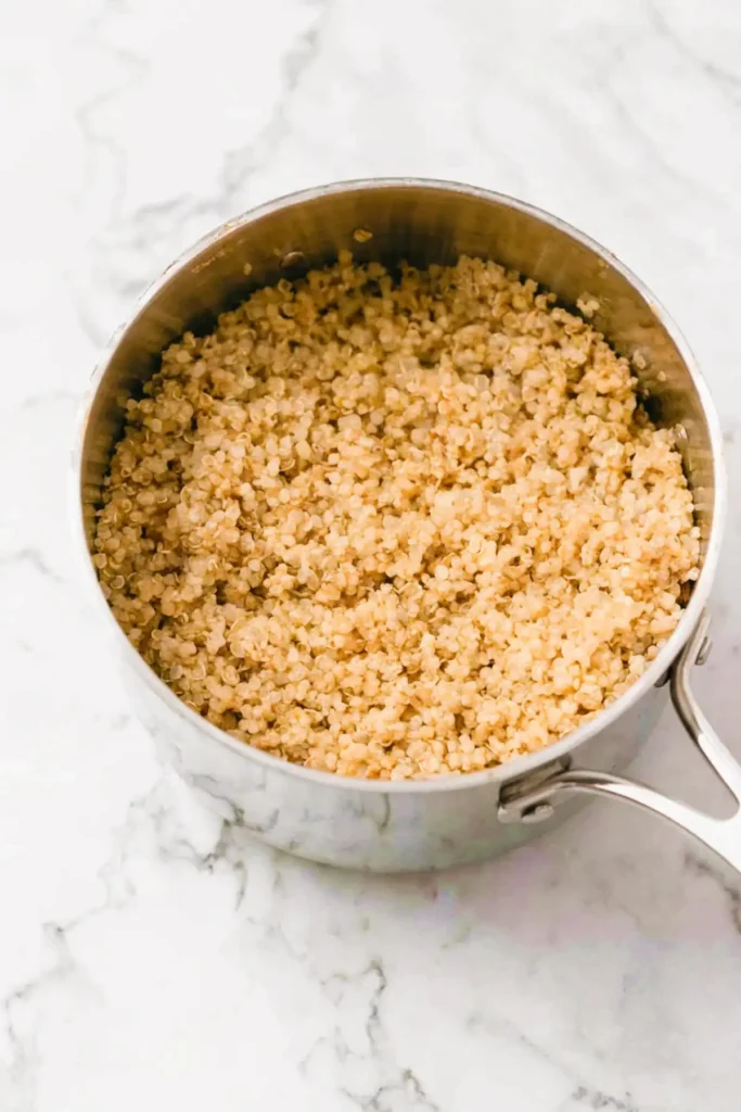 Cooked fluffy quinoa in a stainless steel saucepan on a white marble surface.