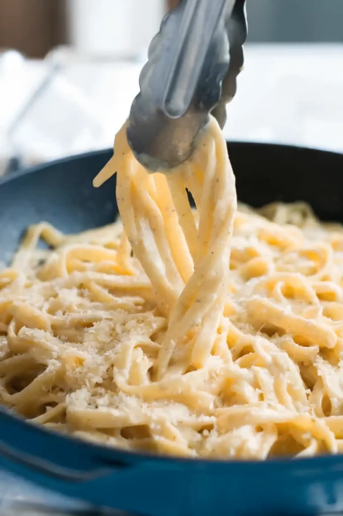 Fettuccine Alfredo being lifted with tongs from a pan with creamy parmesan sauce