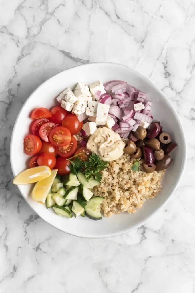 Mediterranean quinoa bowl with cherry tomatoes, cucumber, feta cheese, olives, red onion, hummus, and lemon on a white plate.