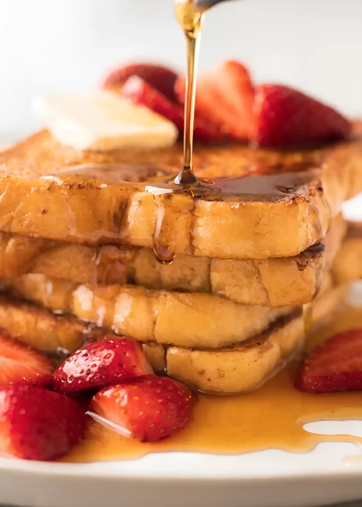 Stack of golden French toast with maple syrup pouring over the top and fresh strawberries on a white plate.