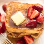 French toast topped with melting butter, maple syrup, and fresh strawberries on a white plate.