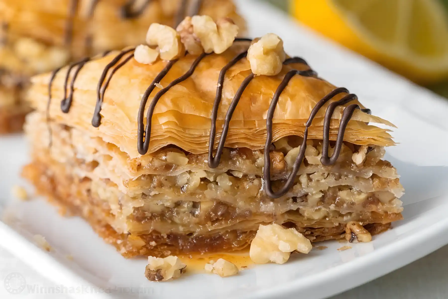 Close up of homemade baklava slice with flaky phyllo layers, walnut filling, honey syrup, and chocolate drizzle on white plate.