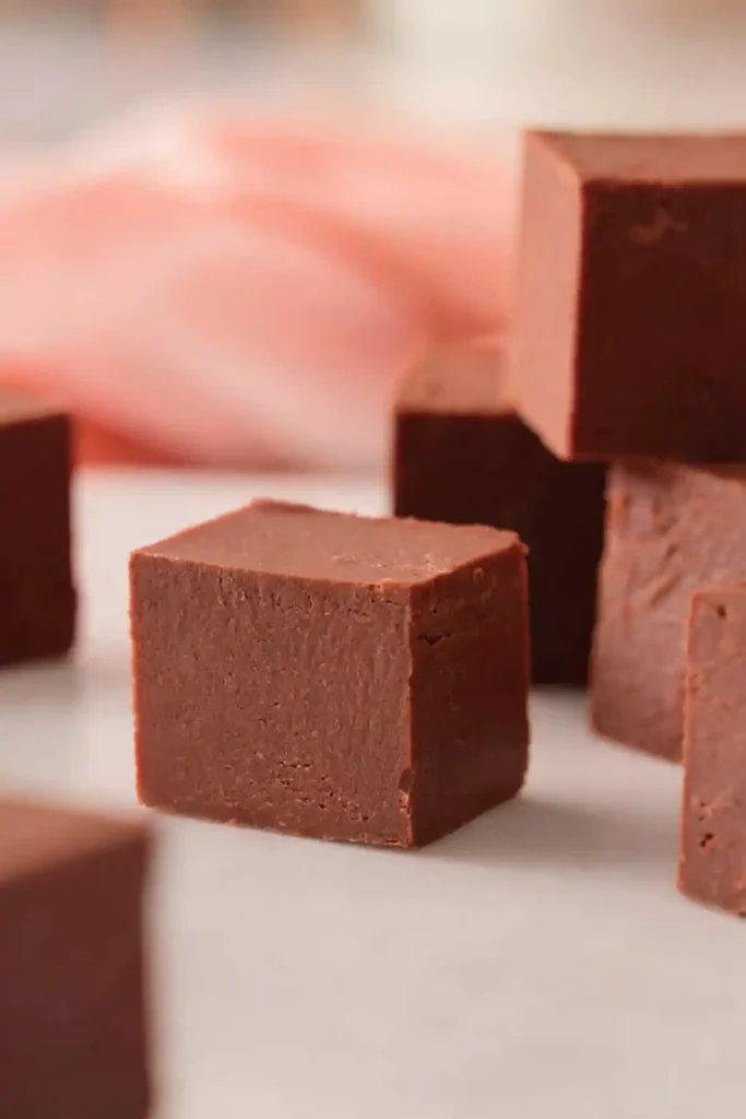 Homemade chocolate fudge recipe cubes with a smooth creamy texture placed on a white surface with softly blurred fudge pieces in the background.