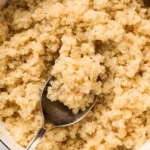 Fluffy cooked quinoa in a white saucepan with a spoon lifting a scoop of light golden quinoa grains.