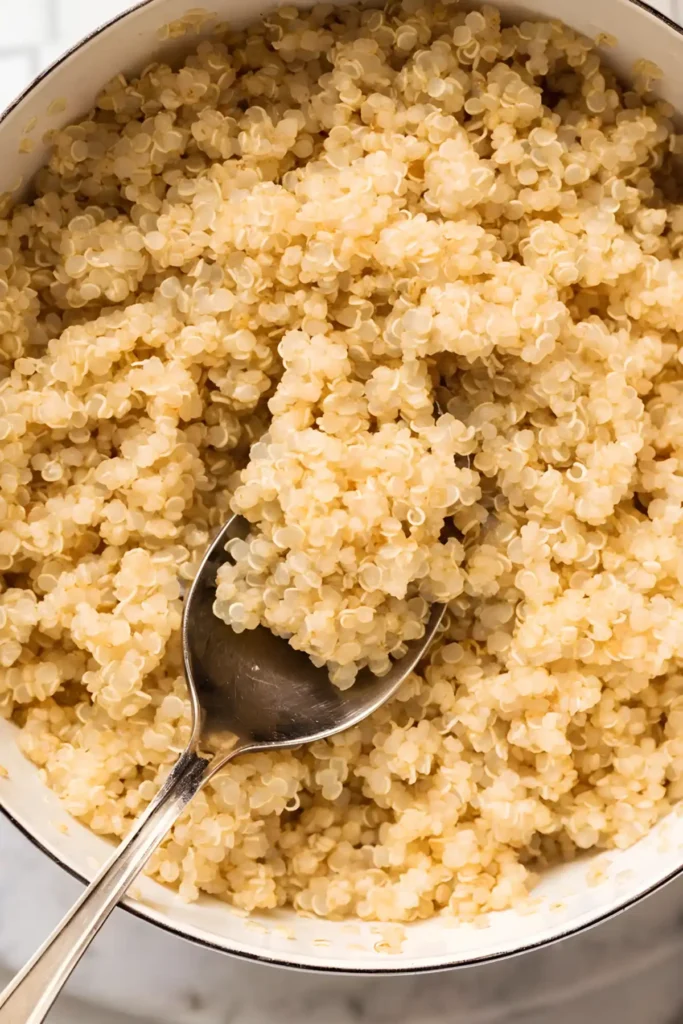 Fluffy cooked quinoa in a white saucepan with a spoon lifting a scoop of light golden quinoa grains.