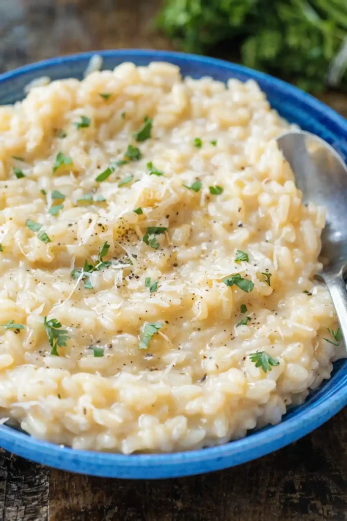Creamy parmesan risotto served in a blue bowl with grated parmesan, chopped parsley, black pepper, and a spoon on the side.