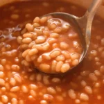 Homemade baked beans in rich tomato sauce with a spoon scooping soft white beans
