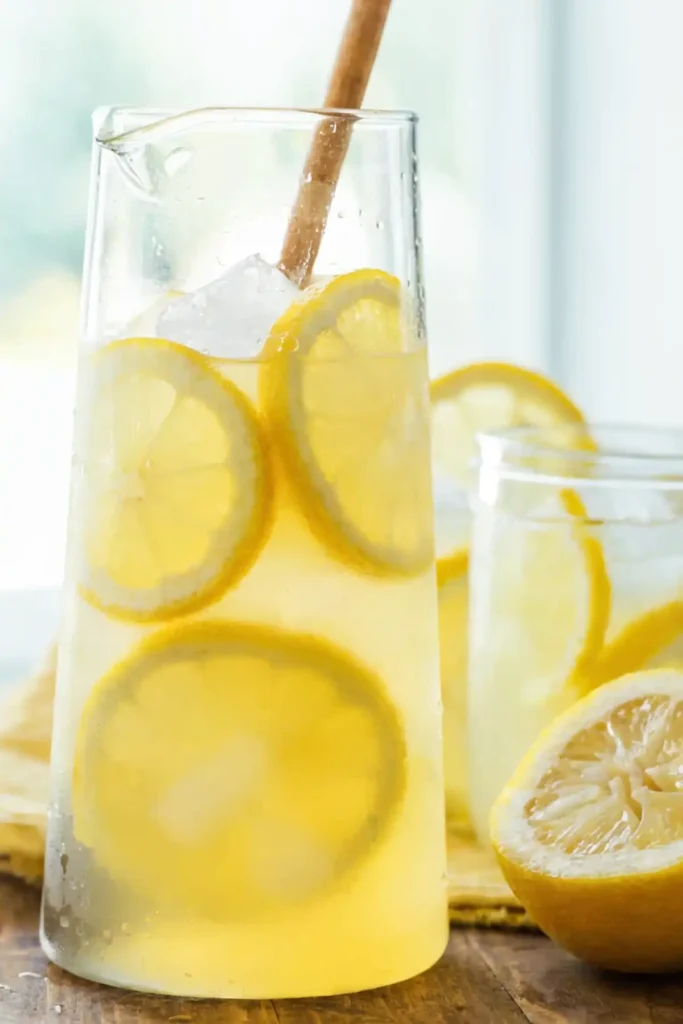 Glass pitcher of fresh homemade lemonade with ice cubes and lemon slices, served on a wooden table with a mason jar and a halved lemon.