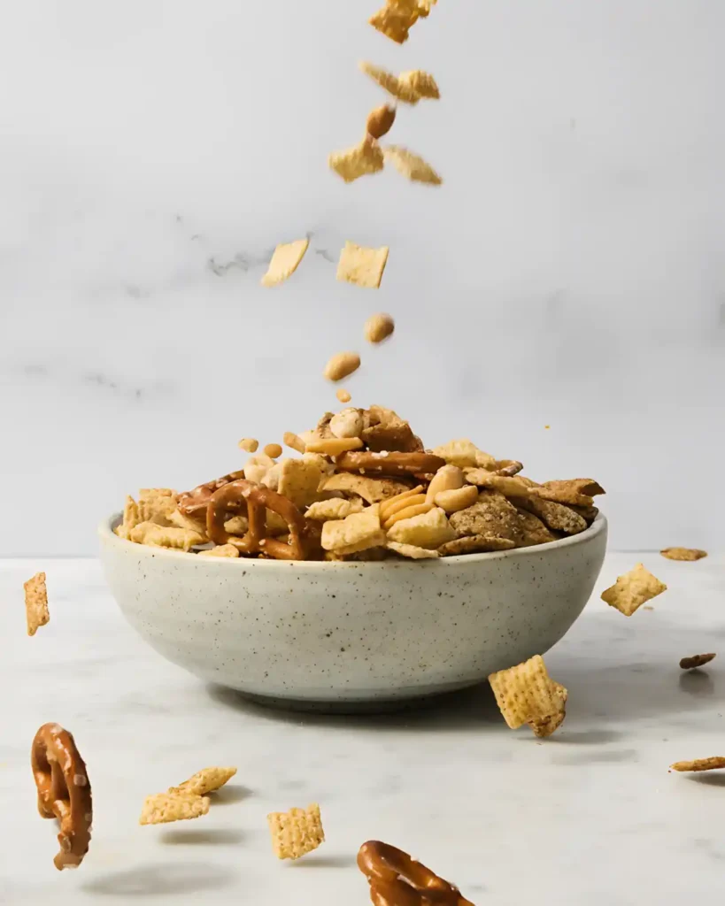 Homemade Chex mix snack falling into a bowl with pretzels, cereal squares, and peanuts.