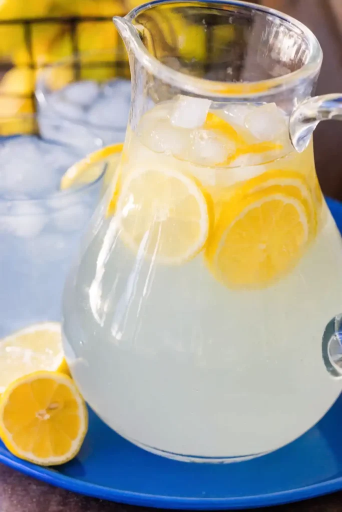 Glass pitcher of homemade lemonade with ice and fresh lemon slices, placed on a blue tray with lemon halves and a glass of lemonade in the background.