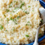 Creamy parmesan risotto served in a blue bowl topped with grated cheese, chopped parsley, and black pepper, with a spoon on the side.