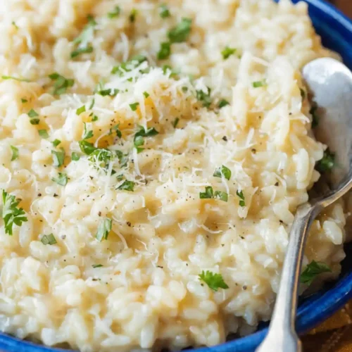 Creamy parmesan risotto served in a blue bowl topped with grated cheese, chopped parsley, and black pepper, with a spoon on the side.