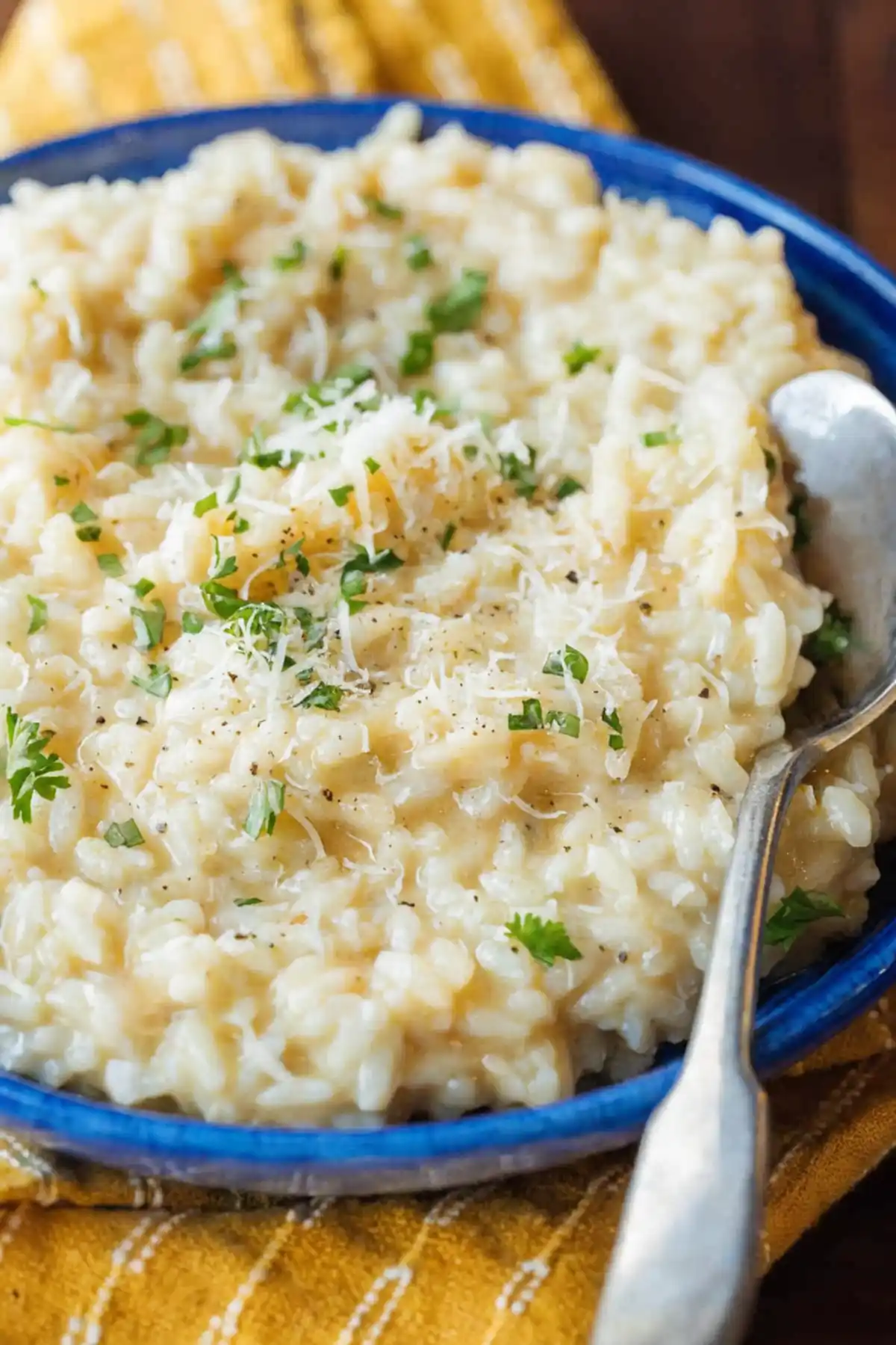 Creamy parmesan risotto served in a blue bowl topped with grated cheese, chopped parsley, and black pepper, with a spoon on the side.