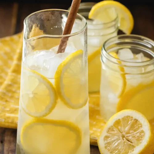 Homemade lemonade in a glass pitcher with ice and fresh lemon slices, served on a wooden table with mason jars and a cut lemon.