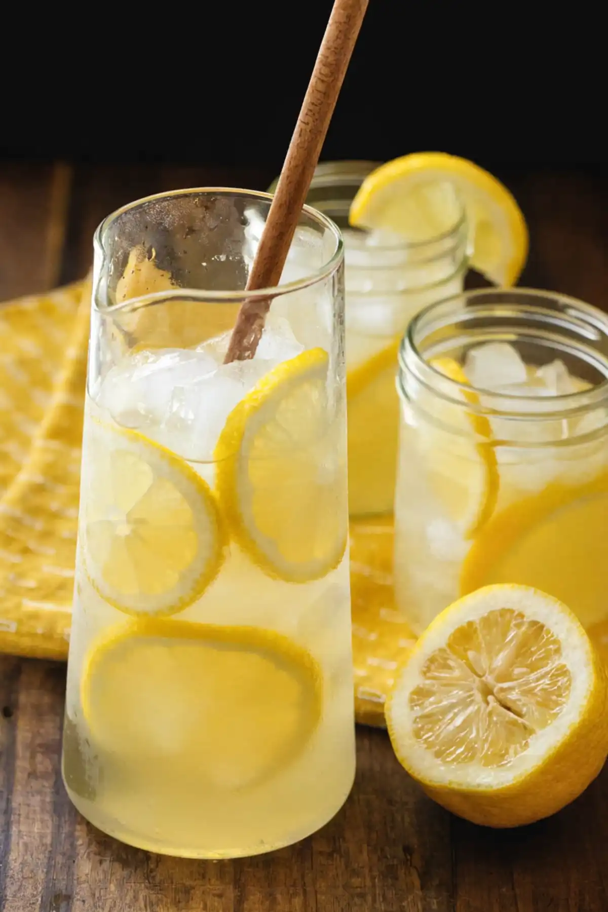 Homemade lemonade in a glass pitcher with ice and fresh lemon slices, served on a wooden table with mason jars and a cut lemon.