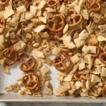 Homemade Chex mix with pretzels, cereal squares, bagel chips, and peanuts spread on a parchment lined baking sheet.