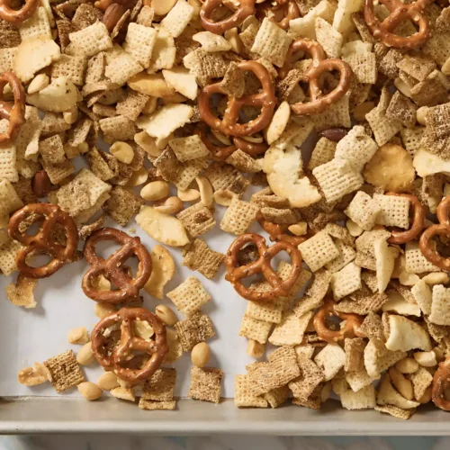 Homemade Chex mix with pretzels, cereal squares, bagel chips, and peanuts spread on a parchment lined baking sheet.