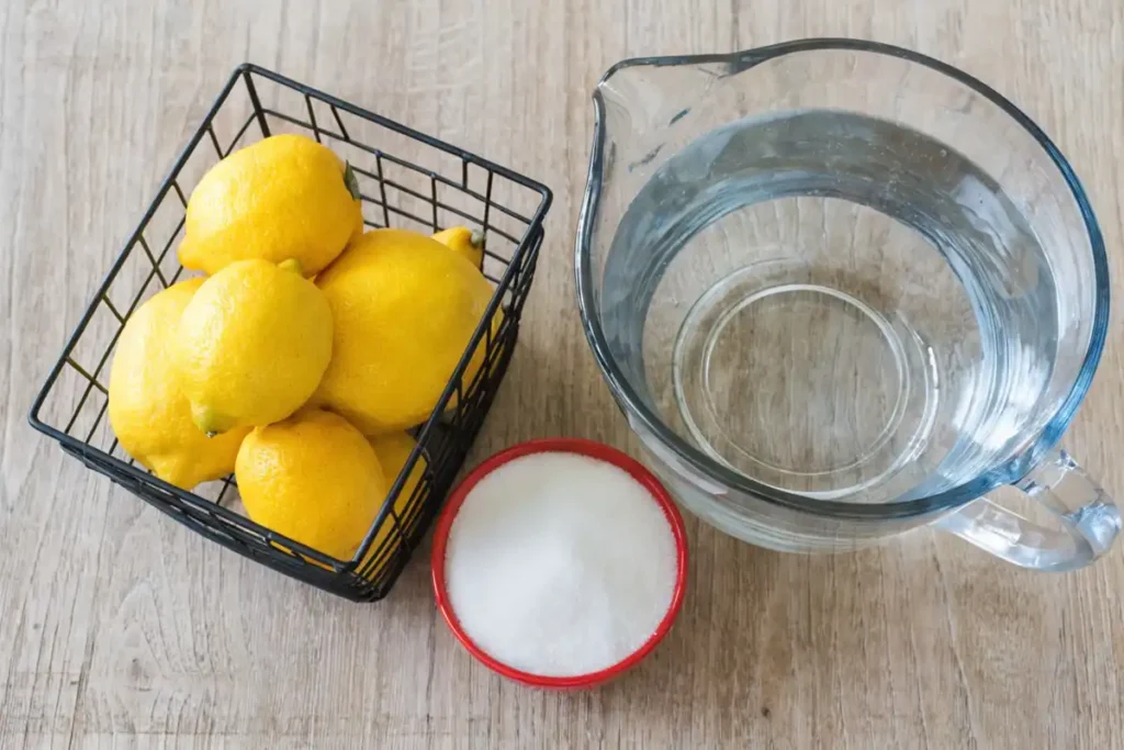 Fresh lemonade ingredients including whole lemons in a basket, a glass pitcher of water, and a bowl of sugar arranged on a wooden table.