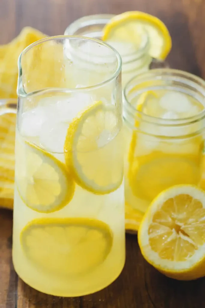 Fresh homemade lemonade in a clear glass pitcher with ice cubes and lemon slices, served on a wooden table with mason jars and a halved lemon.