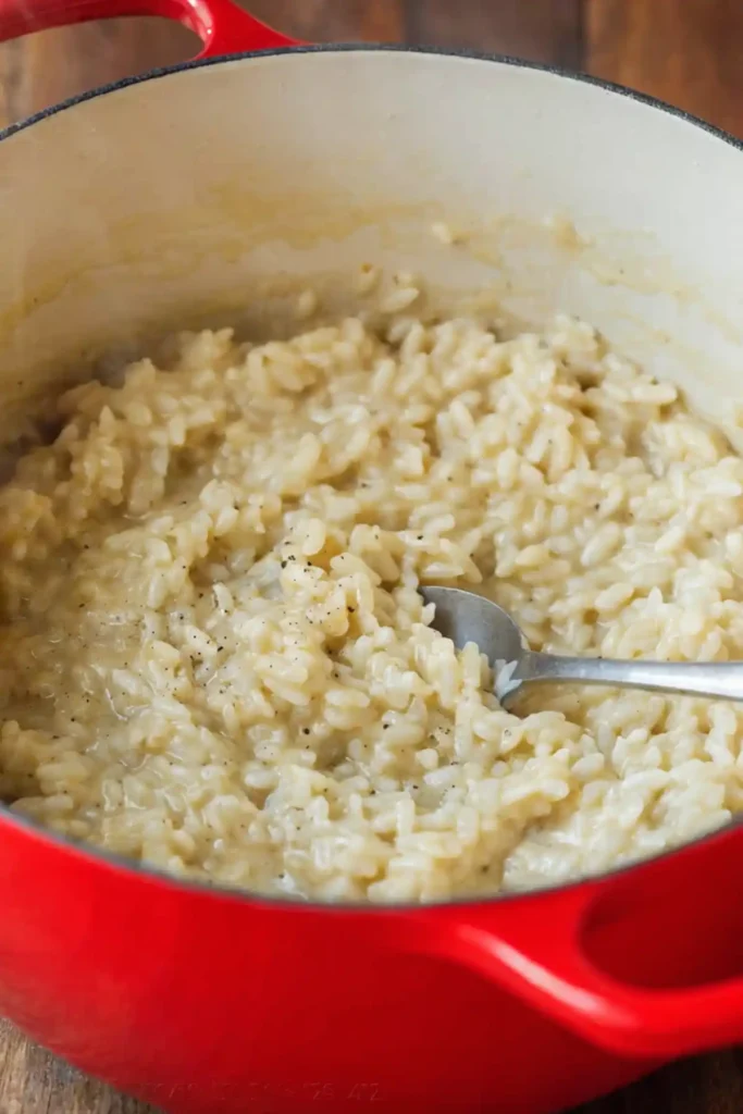 Creamy risotto cooking in a red Dutch oven with Arborio rice and a spoon stirring the rich, glossy mixture.