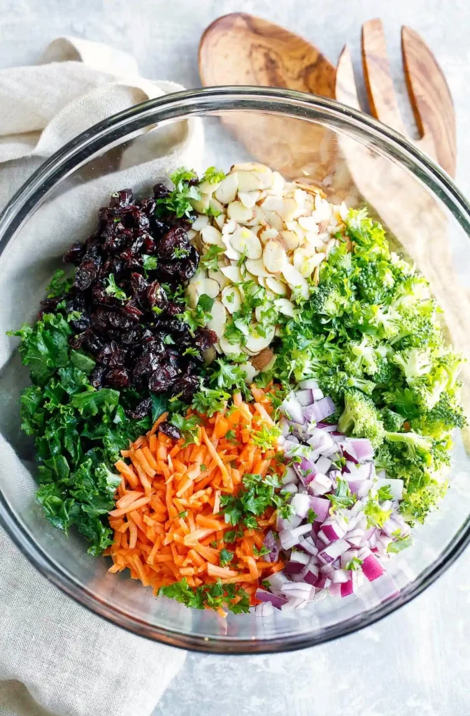Kale salad ingredients in a glass bowl with chopped kale, broccoli, shredded carrots, red onion, sliced almonds, and dried cranberries before mixing.