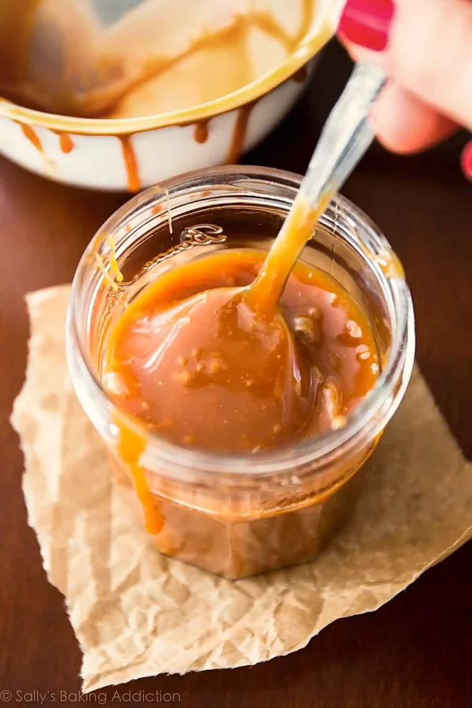 Homemade caramel recipe in a jar being stirred with a spoon showing thick texture and small sugar bits