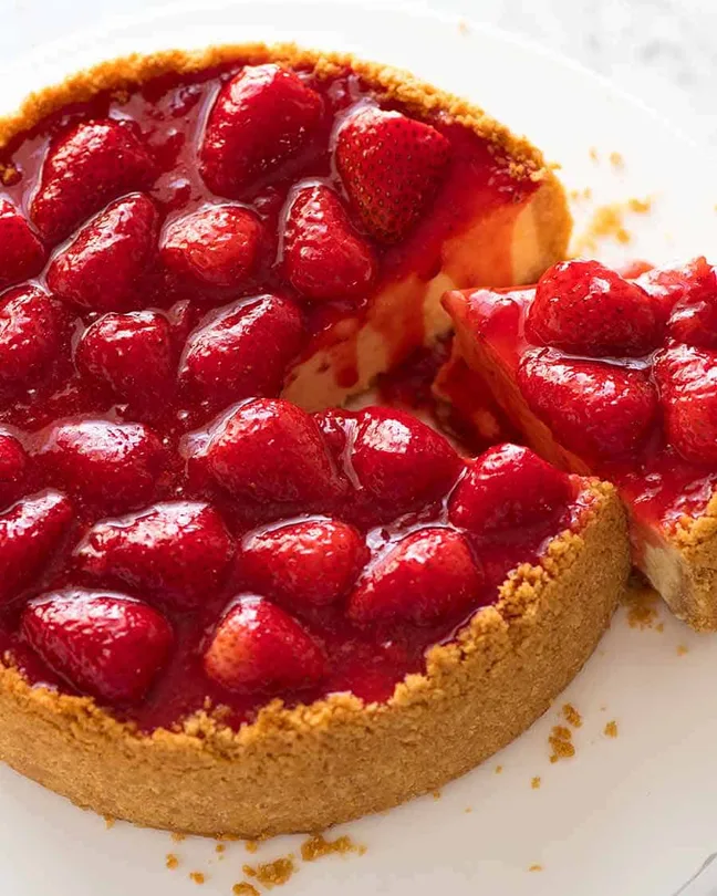 Whole strawberry cheesecake with a slice removed, topped with fresh strawberries and glossy red sauce on a golden biscuit crust