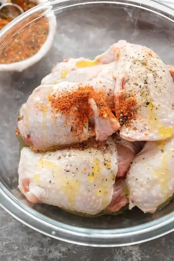 Raw chicken thighs in a bowl with olive oil and seasoning including paprika, garlic, and herbs