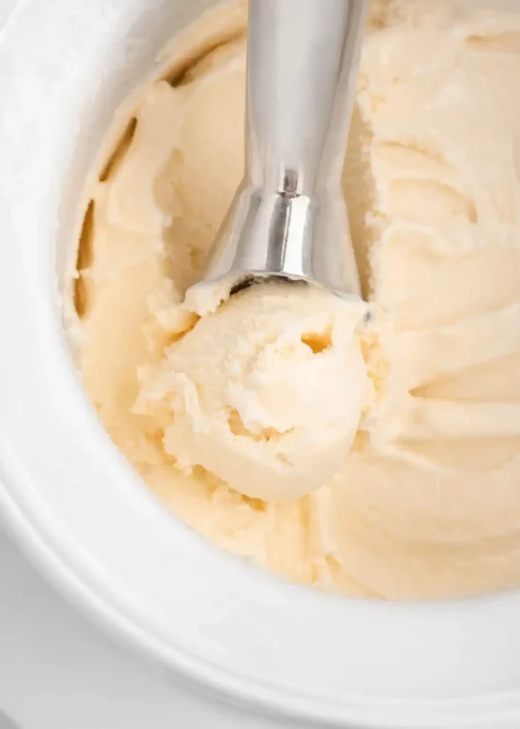 Ice cream scoop lifting creamy homemade vanilla ice cream from a white container on a bright background.