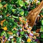 Fresh kale salad with broccoli, shredded carrots, red onion, sunflower seeds, dried cranberries, and cheese, tossed in a light lemon dressing with a wooden spoon.