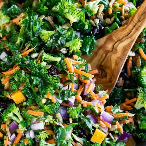 Fresh kale salad with broccoli, shredded carrots, red onion, sunflower seeds, dried cranberries, and cheese, tossed in a light lemon dressing with a wooden spoon.