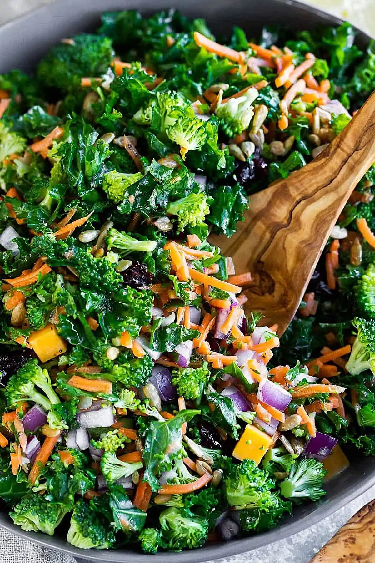 Fresh kale salad with broccoli, shredded carrots, red onion, sunflower seeds, dried cranberries, and cheese, tossed in a light lemon dressing with a wooden spoon.