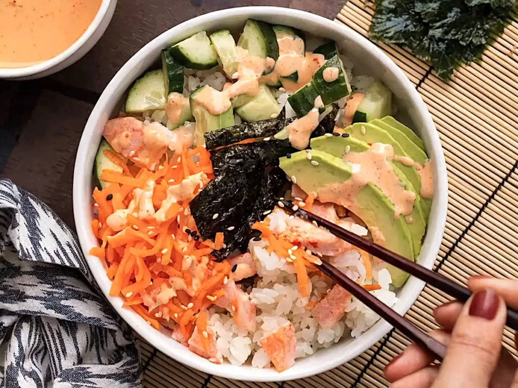 Sushi bowl with crab, avocado, cucumber, carrot, sesame seeds, and spicy mayo with chopsticks picking a bite
