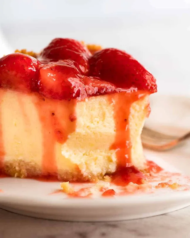 Close up of strawberry cheesecake slice with creamy filling and fresh strawberry topping dripping with glossy red sauce