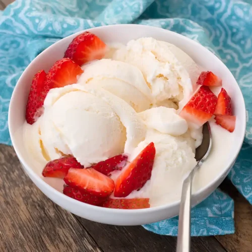 Bowl of homemade vanilla ice cream topped with fresh sliced strawberries on a wooden table with a blue cloth background.