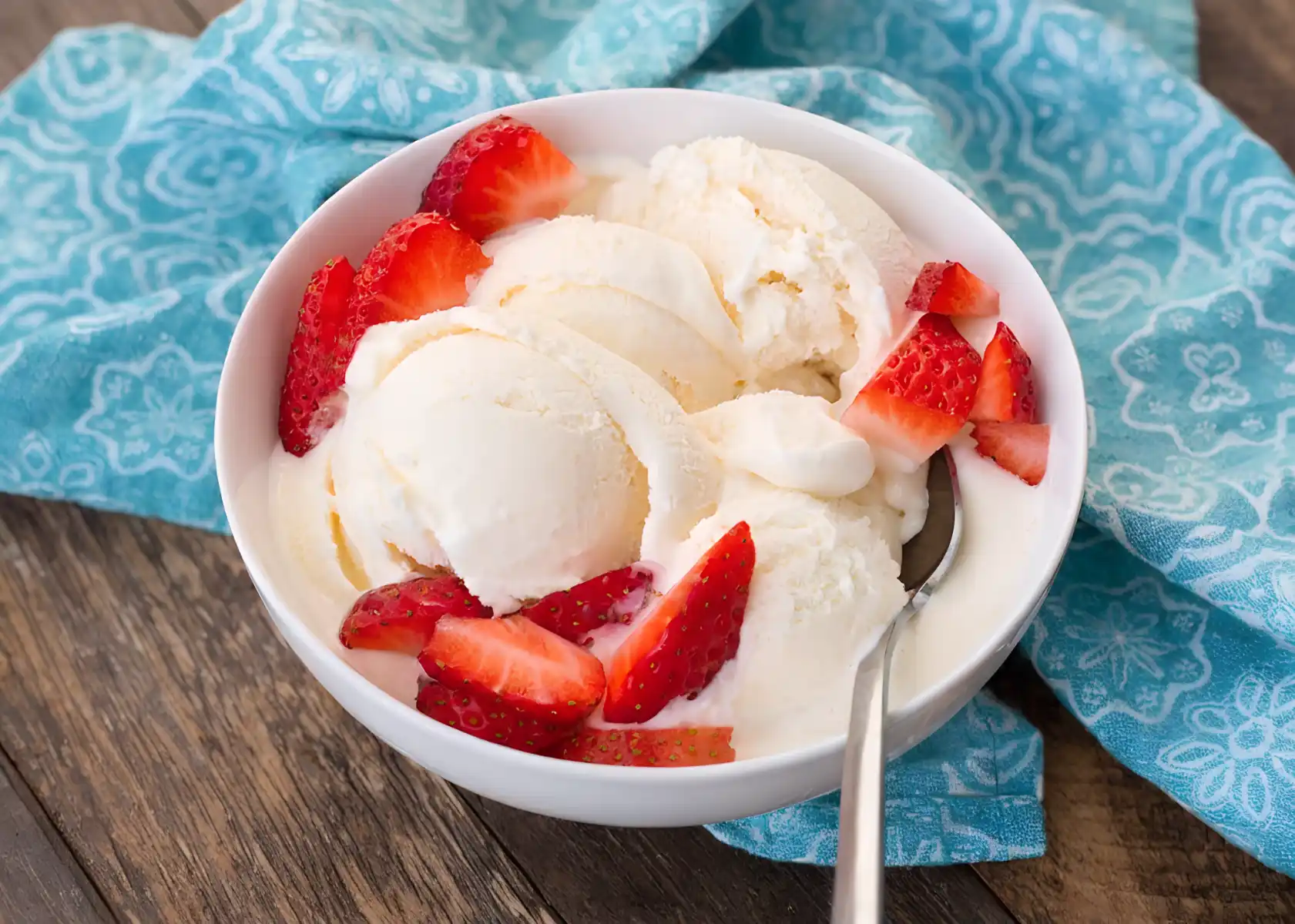 Bowl of homemade vanilla ice cream topped with fresh sliced strawberries on a wooden table with a blue cloth background.