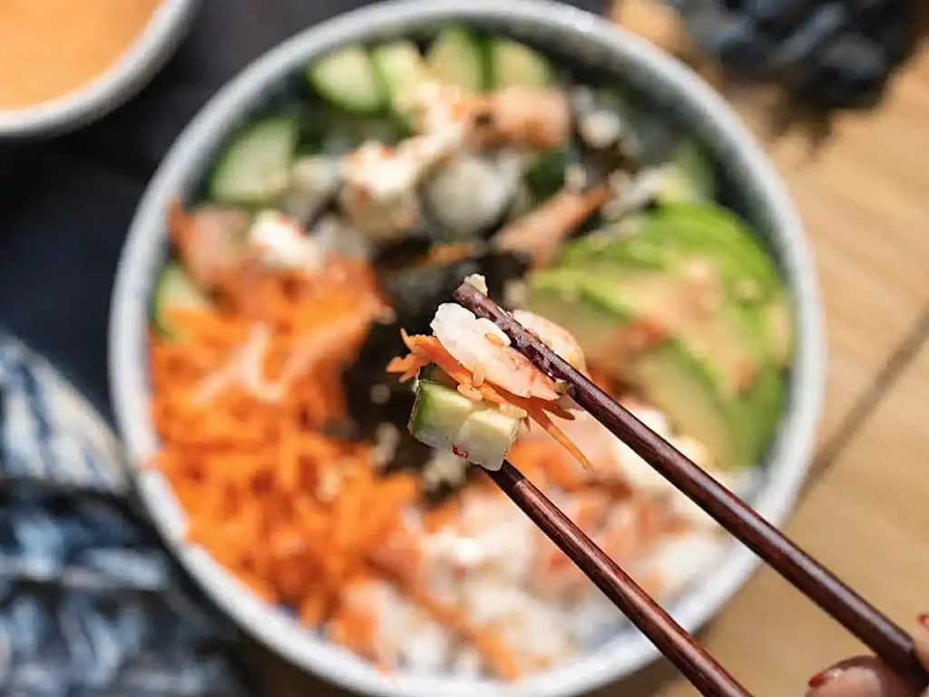 Homemade sushi bowl with chopsticks lifting a bite of crab, rice, and vegetables with spicy mayo
