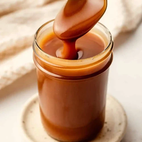 Homemade caramel sauce dripping from a spoon into a glass jar with smooth glossy texture