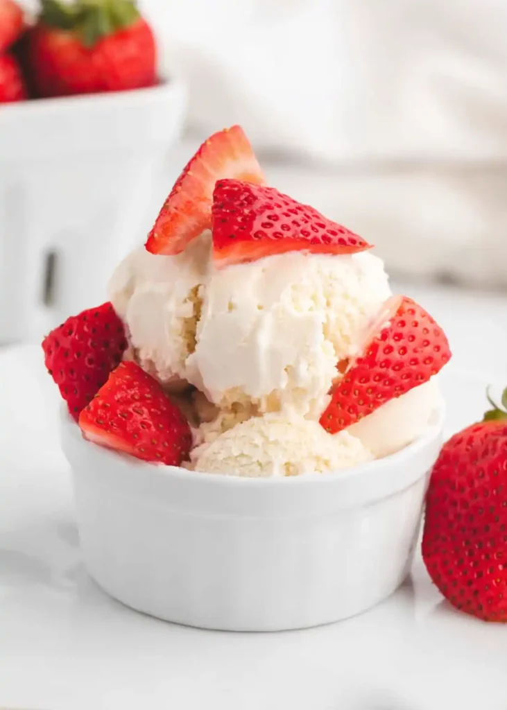 Small ramekin of homemade vanilla ice cream topped with fresh sliced strawberries on a bright white surface.