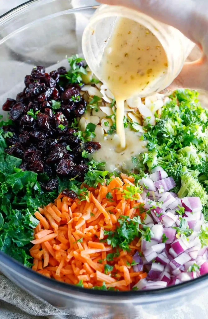 Lemon dressing being poured over kale salad with broccoli, shredded carrots, red onion, almonds, and dried cranberries in a mixing bowl.