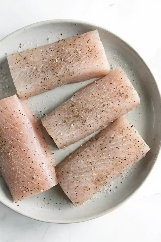 Raw mahi mahi fillets on a plate, seasoned with salt and black pepper before cooking.