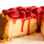 Close-up of creamy strawberry cheesecake slice with fresh strawberry topping and glossy red sauce dripping over a golden biscuit crust