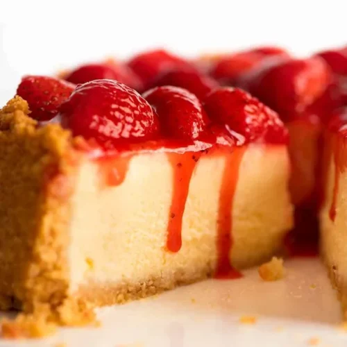 Close-up of creamy strawberry cheesecake slice with fresh strawberry topping and glossy red sauce dripping over a golden biscuit crust