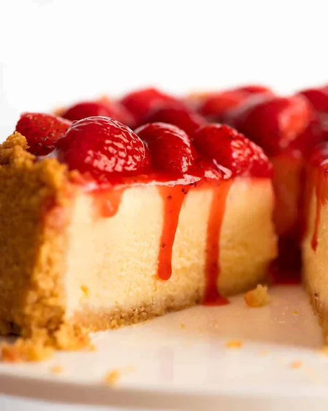 Close-up of creamy strawberry cheesecake slice with fresh strawberry topping and glossy red sauce dripping over a golden biscuit crust